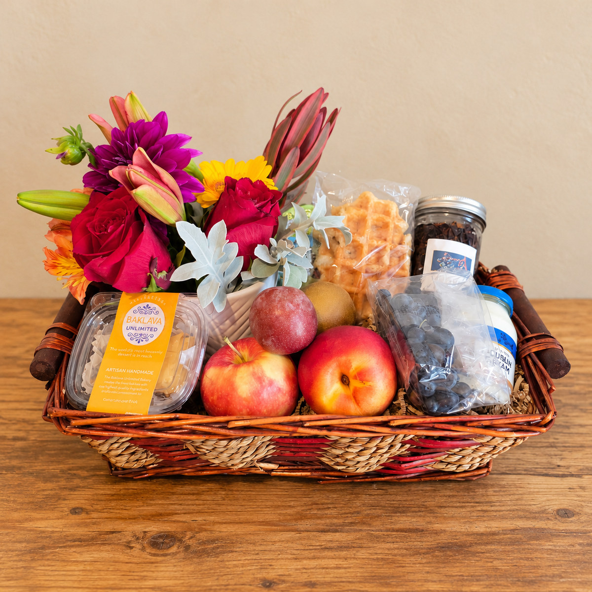 Wicker gift basket combining a floral arrangement, fresh fruit, pastries, chocolate, and other treats